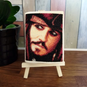 Puede incluir: Un retrato de punto de cruz de un hombre con barba y bigote oscuros, que lleva un sombrero rojo. El retrato está enmarcado y se muestra en un pequeño caballete de madera.