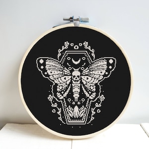 Puede incluir: Aro de bordado en blanco y negro con un diseño de punto de cruz de una polilla con una calavera en su cuerpo, dentro de un ataúd con una luna creciente y estrellas arriba.