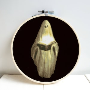 Puede incluir: Un fantasma blanco con un libro en sus manos, bordado sobre un fondo negro en un aro de bordado de madera redondo.