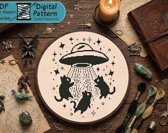 May include: A circular embroidery hoop containing a cross-stitch pattern of a black UFO hovering above three black cats, with a beam of light shining down on them, surrounded by small stars on a light beige fabric background.