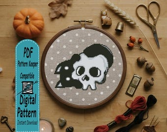 May include: A cross-stitch project in a wooden hoop featuring a white skull with black hair on a beige fabric with white polka dots. A blue vertical banner on the left displays the text PDF Pattern Keeper Compatible Digital Pattern.