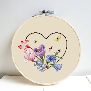 Peut inclure: Un cerceau de broderie au point de croix avec un motif en forme de cœur représentant une variété de fleurs dans des tons de rose, violet et bleu. Les fleurs sont entourées d'abeilles et de papillons.