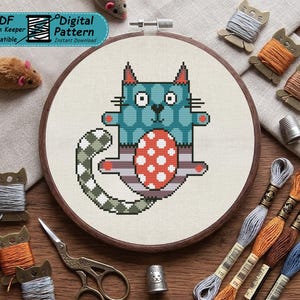 Peut inclure: Un motif de point de croix dans un tambour en bois représentant un chat bleu sarcelle avec un ventre à pois rouges et blancs et une queue à carreaux verts et blancs. Le design inclut une étiquette PDF Pattern Keeper Compatible, du fil, des ciseaux et des dés.