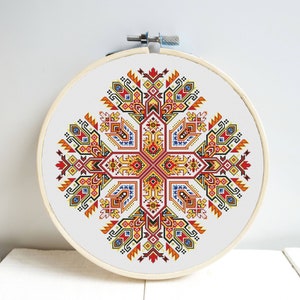 May include: A cross-stitch embroidery featuring a symmetrical geometric design in red, yellow, blue, and orange hues. The artwork is displayed in a round wooden embroidery hoop, showcasing a traditional craft.