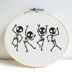 May include: A black and white cross-stitch pattern of four skeletons dancing. The skeletons are in a circle, with their arms and legs outstretched. The pattern is on a white background.