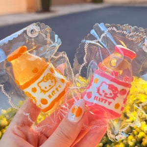 Puede incluir: Dos pequeños llaveros con forma de botella de leche coloridas. Uno es naranja y el otro rosa, ambos con un diseño de Hello Kitty. Están empaquetados en plástico transparente en forma de corazón con una cadena plateada.
