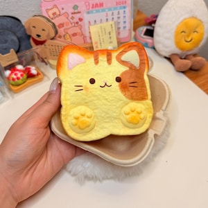 May include: A soft, squishy foam toy shaped like a slice of toast with a cat face. The toast is yellow with brown patches, featuring two cat ears, a small nose, whiskers, and two raised paw prints at the bottom, all held in a small paper container.