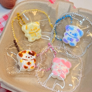 May include: A collection of colorful dog-shaped keychains in a cardboard container. The keychains are in clear plastic pouches and attached to beaded chains. The dogs are yellow, blue, brown, and pink with various patterns. The container has a pink and white checkered background.
