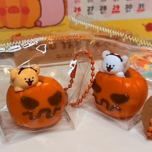 May include: Two Halloween-themed keychains. Each keychain features a small bear figurine sitting inside an orange pumpkin. The pumpkins have carved faces. The keychains are in clear plastic bags, with a calendar in the background.