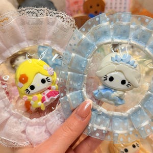 May include: Two Hello Kitty character ornaments, one yellow and one blue, are displayed in clear packaging. Each is surrounded by a ruffled lace collar. The yellow ornament has a pink dress, while the blue one wears a blue dress.