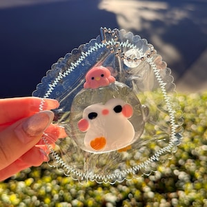 May include: Clear acrylic keychain featuring a cartoon penguin with a pink octopus on top. The penguin has black eyes, pink cheeks, and an orange belly. The keychain has a scalloped edge and a silver chain.