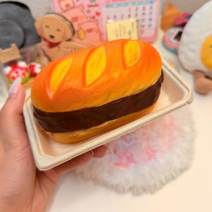 May include: A squishy toy shaped like a baguette sandwich with a golden-brown crust featuring three diagonal slashes on top and a dark brown filling layer, presented in a light beige rectangular paper tray.