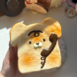 May include: A square-shaped piece of toasted bread-shaped decor featuring a light brown toasted surface with a cartoon bear face. The bear has a dark brown telephone handset graphic on the right side and a small dark brown curl on its forehead.