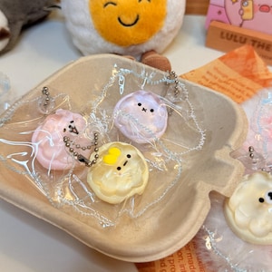 May include: A collection of pastel-colored, round keychains with small animal faces, packaged in clear plastic. A plush egg with a smiling face and a calendar with pig illustrations are in the background. The keychains are in a small, light brown container.