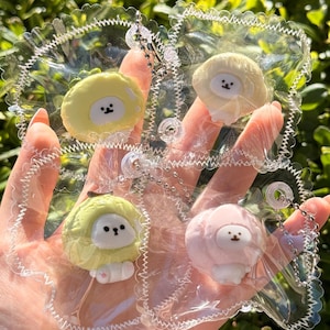 May include: Four mushroom-shaped keychains in yellow, cream, green, and pink, each with a small, white face. The keychains are attached to clear, scalloped-edged plastic holders with silver chains. The background is blurred green foliage.