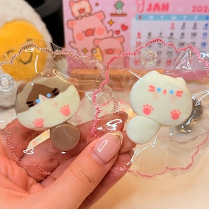 Könnte beinhalten: Zwei katzenförmige Schlüsselanhänger. Einer ist weiß mit rosa Pfoten und blauen Akzenten, der andere ist braun und cremefarben mit rosa Pfoten. Beide befinden sich in klarem Kunststoff mit rosa Besatz. Im Hintergrund befindet sich ein Kalender mit Schweineillustrationen.