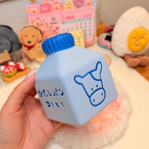 May include: A light blue square-shaped squishy toy designed to resemble a milk carton. It features a dark blue screw-top cap, a graphic of a cow face, and Japanese text printed in dark blue ink on the front side of the carton.