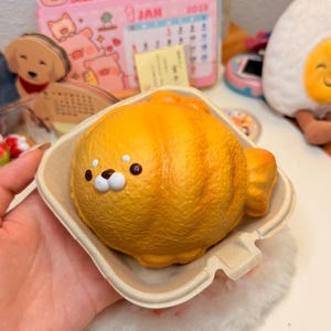 May include: A golden-yellow, fish-shaped squishy toy with a textured surface, featuring a small white snout, two dark brown eyes, and two small white highlights above the eyes, presented inside a light beige, square-shaped, molded fiber container.