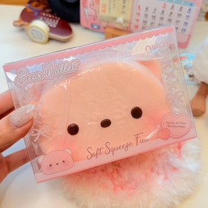 May include: A light pink bear-shaped squeeze toy wallet inside a clear plastic retail box. The packaging features the text Bear Wallet and Soft Squeeze Fun, with a small heart graphic that reads Squish All Your Worries Away. The toy has two dark brown eyes and a small dark brown nose.