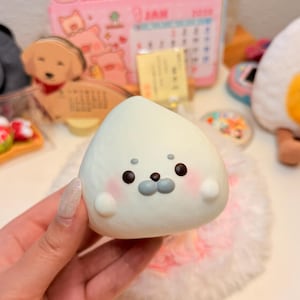 May include: A white, rounded, squishy seal-shaped toy with a smooth texture. It features two small black eyes, two grey dots above the eyes, a grey oval nose, and soft pink blush marks on its cheeks. The toy has two small, rounded side fins.
