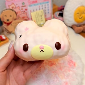 May include: A light pink squishy toy shaped like a bear head with a yellow muzzle, dark brown eyes, and a small pink nose. The bear features two small star-shaped prints on the upper left side and soft, rounded ears on top of its head.