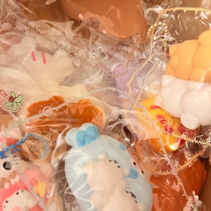 Puede incluir: Una colección de jabones hechos a mano de diversas formas, incluyendo osos y donuts. Los jabones están envueltos individualmente en plástico transparente, creando una presentación similar a un regalo. La paleta de colores es suave y pastel.