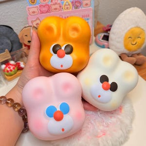May include: Three squishy toys shaped like animal heads with rounded ears. One is orange with dark brown eyes and a red nose, one is white with dark brown eyes and a red nose, and one is light pink with blue eyes and a red nose on a white muzzle.