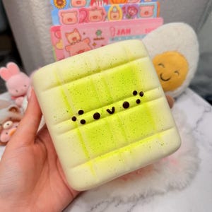 May include: A square, cream-colored squishy toy with a grid pattern and a bright neon green spray-painted effect. The center features a simple, dark brown dotted face with a small v-shaped mouth, resembling a stylized character expression.
