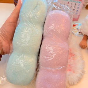 May include: Two elongated, cylindrical plush items with a soft, textured surface, each individually wrapped in clear plastic. One item is light blue and the other is light pink, both featuring a segmented, rounded shape resembling a long pillow.