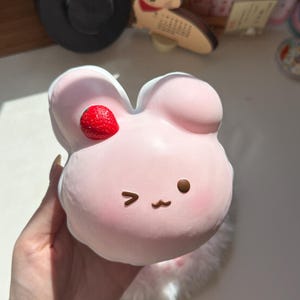 May include: A soft, light pink, rounded squishy toy shaped like a rabbit head. It features a small, textured red strawberry attached to the right ear, a winking eye, a circular eye, and a simple brown mouth. The surface has a smooth, matte finish.