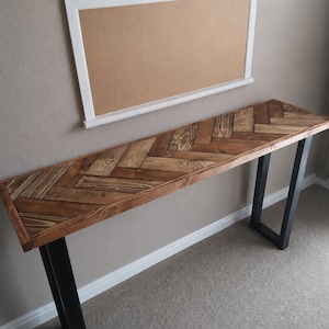 May include: A long, narrow console table featuring a rectangular top made of dark brown stained wood planks arranged in a herringbone pattern. The table is supported by two black metal U-shaped legs. Above the table hangs a rectangular cork board.