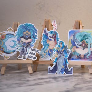 Peut inclure: Plusieurs autocollants représentant un personnage aux cheveux bleus et un masque. Ils sont présentés sur de petits chevalets en bois. L'un des autocollants porte l'inscription "I don't say that to flatter you."