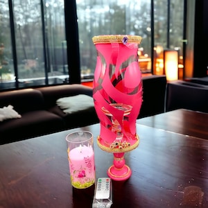 Puede incluir: Un portavelas de vidrio rosa con detalles dorados y un diseño decorativo. El portavelas está sobre una mesa de madera con una vela rosa en un frasco de vidrio.