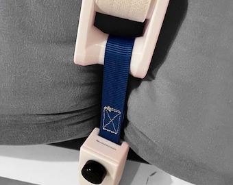 May include: A light pink plastic tensioning device with a white fabric strap and a blue ribbon strap. The device features a black circular knob and is positioned between two gray fabric cushions, securing them together with its mechanical components.