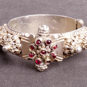 Puede incluir: Un brazalete de plata con intrincados detalles de filigrana y un diseño central con un grupo de piedras preciosas rojas.