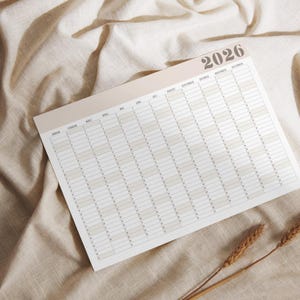May include: A yearly planner for 2026, featuring a neutral color palette. The calendar is laid out horizontally with monthly and daily sections. The planner is on a beige fabric surface, with a few sprigs of wheat in the lower right corner.