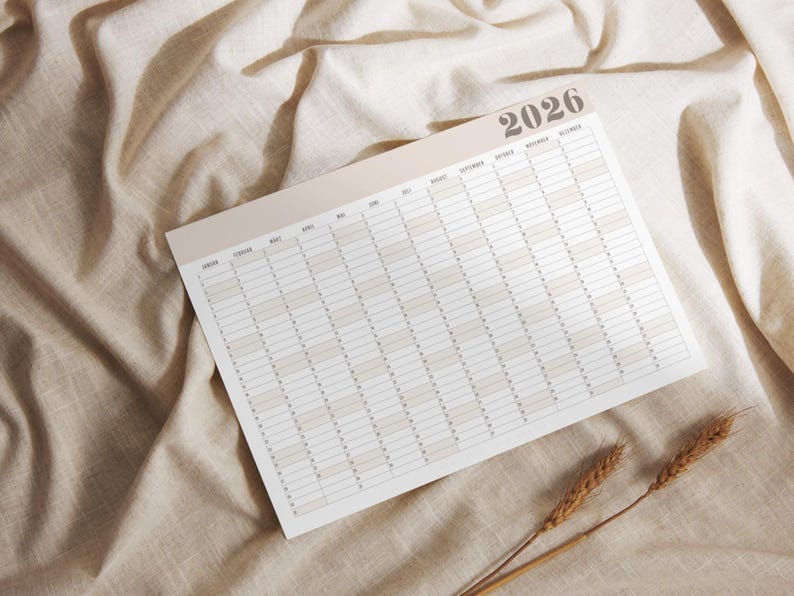 May include: A yearly planner for 2026, featuring a neutral color palette. The calendar is laid out horizontally with monthly and daily sections. The planner is on a beige fabric surface, with a few sprigs of wheat in the lower right corner.