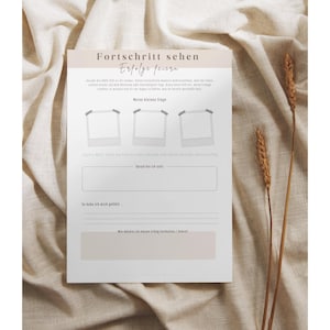 May include: A white journal page with the German text "Fortschritt sehen, Erfolge feiern" and space for notes. The page has three photo placeholders and lines for writing. Two stalks of wheat are on the right side.