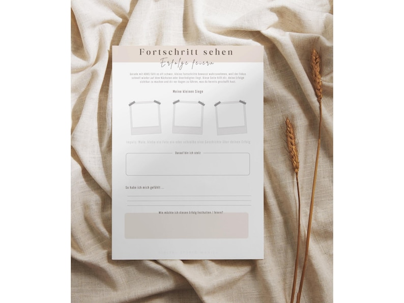 May include: A white journal page with the German text "Fortschritt sehen, Erfolge feiern" and space for notes. The page has three photo placeholders and lines for writing. Two stalks of wheat are on the right side.