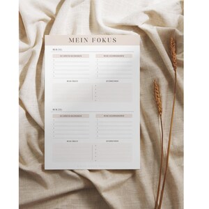 May include: A printable planner page with the title "Mein Fokus" in German. The page is divided into four sections, each with the heading "Mein Ziel" and space for writing down goals and action steps. The page is designed in a minimalist style with a light beige background.
