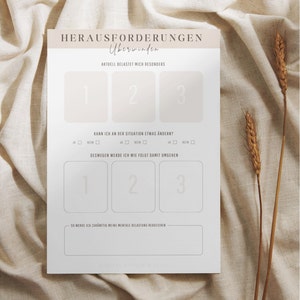 May include: A white paper with German text, including "HERAUSFORDERUNGEN &Uuml;berwinden" and numbered sections for reflection. The paper is on a beige fabric background, with dried wheat stalks to the right.