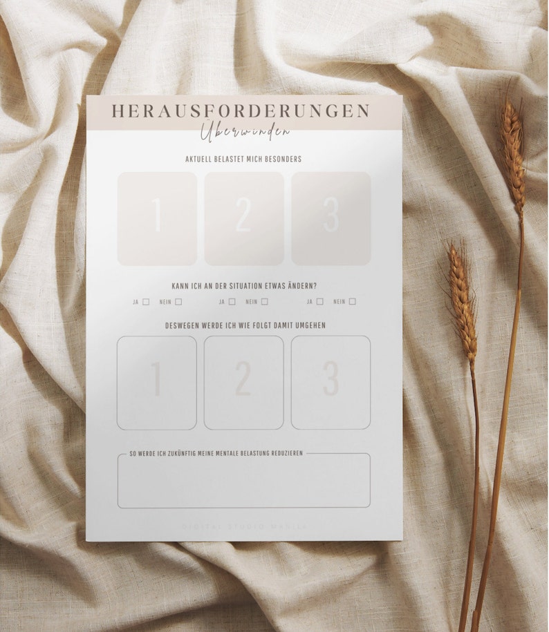 May include: A white paper with German text, including "HERAUSFORDERUNGEN &Uuml;berwinden" and numbered sections for reflection. The paper is on a beige fabric background, with dried wheat stalks to the right.