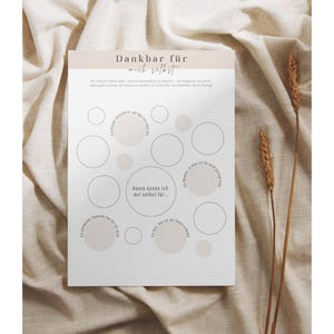 May include: A white paper with the German text "Dankbar f&uuml;r mich selbst" and a series of circles. The paper is on a beige fabric background with two stalks of wheat. The circles are in various sizes and some are filled with a light beige color.