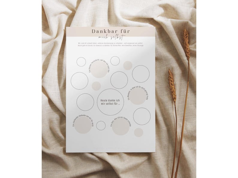 May include: A white paper with the German text "Dankbar f&uuml;r mich selbst" and a series of circles. The paper is on a beige fabric background with two stalks of wheat. The circles are in various sizes and some are filled with a light beige color.