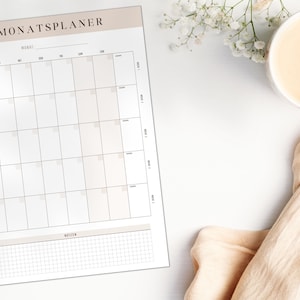 May include: A monthly planner with the German title "MONATSPLANER" at the top, featuring a grid layout for daily scheduling and a notes section. A cup of coffee and a peach-coloured cloth are on the white surface. Measurements are in cm.