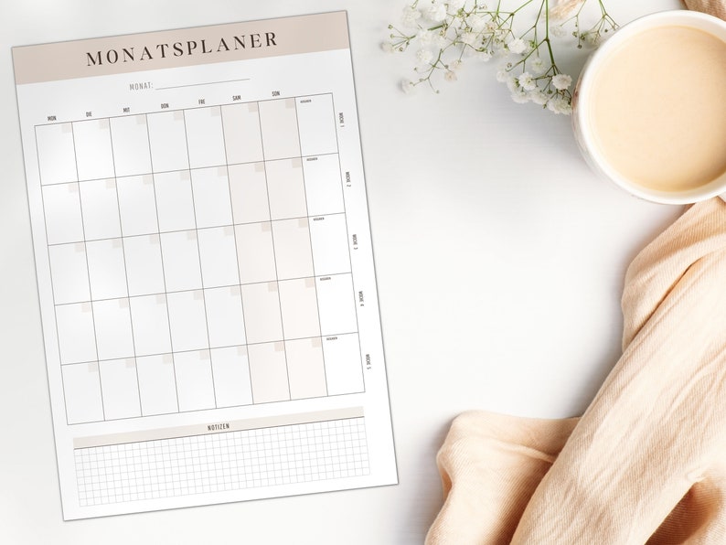 May include: A monthly planner with the German title "MONATSPLANER" at the top, featuring a grid layout for daily scheduling and a notes section. A cup of coffee and a peach-coloured cloth are on the white surface. Measurements are in cm.