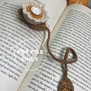 May include: A crocheted bookmark with a brown and beige flower design. The bookmark is shaped like a leaf and has a brown and beige crocheted tassel. The bookmark is lying on top of an open book with the text "Cinderella" visible on the page.