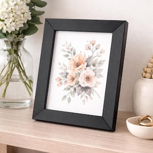 Peut inclure: Un cadre noir contenant une impression florale à l'aquarelle aux tons pêche et blancs doux, avec des feuilles gris-vert. L'art encadré est posé sur une étagère en bois clair, avec un vase de fleurs blanches et de verdure.