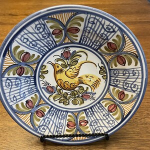 May include: A white ceramic plate with a blue and yellow floral design. The plate features a stylized bird with a yellow body and blue wings. The plate has a decorative border with blue and yellow stripes.