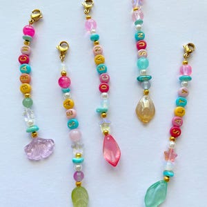 Peut inclure: Cinq breloques perlées avec fermoirs dorés. Chaque breloque présente des perles colorées épelant des mots comme "BEACH", "SEA", "OCEAN", "SUN" et "SALTY". Les breloques comprennent des éléments décoratifs tels qu'un coquillage, une feuille et des pendentifs en forme de goutte.